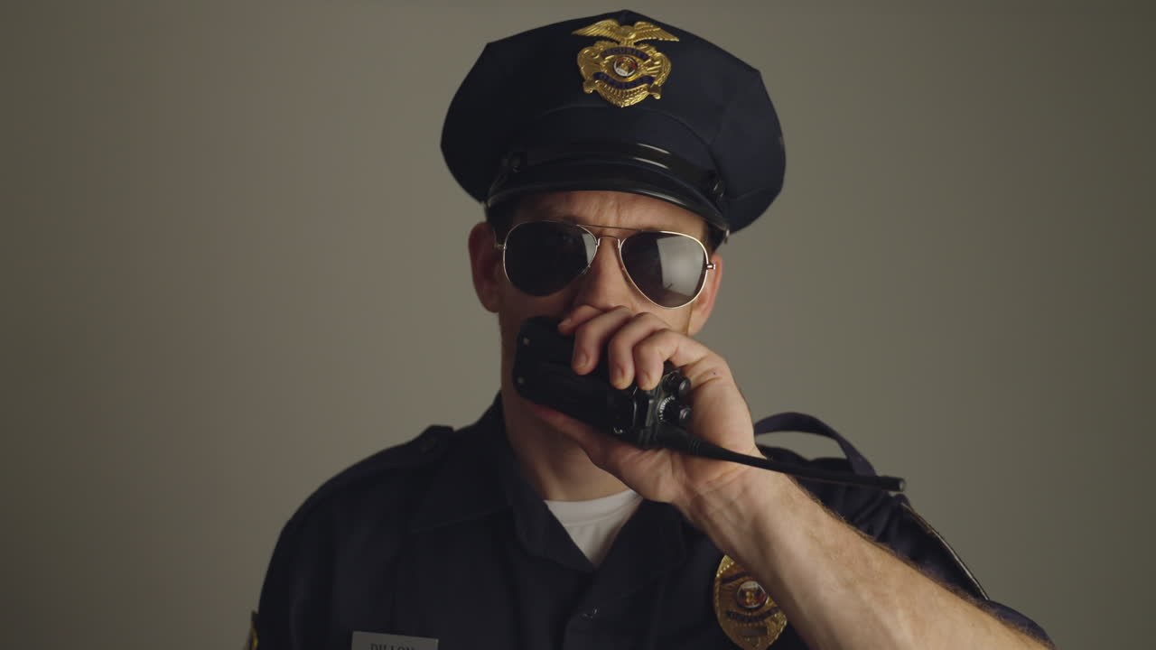 Police officer or cop portrait with sunglasses and red and blue lights flashing. Policeman with blue uniform looking at camera on gray background talking on radio dispatch and looking serious