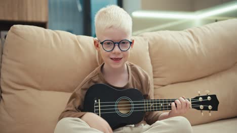 retrato de un feliz niño albino con el color del cabello blanco en gafas redondas azules que toca un ukulele negro y se sienta en un sofá de crema en un apartamento moderno en casa