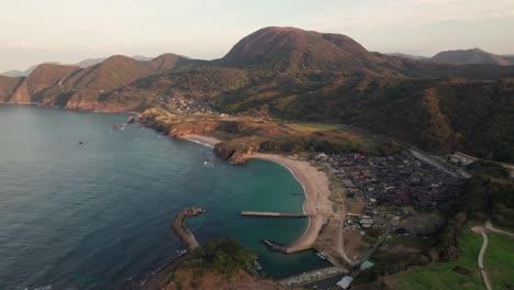 Paisaje-Aéreo-De-Drones-De-La-Bahía-Japonesa-Del-Mar-Azul-En-La-Costa-De-Verano-De-La-Isla-De-Kyoto-Kyotango,-Playa-De-Arena-Blanca-Con-Un-Pueblo-Cercano,-Japón-Durante-El-Día,-Fondo-De-La-Cordillera