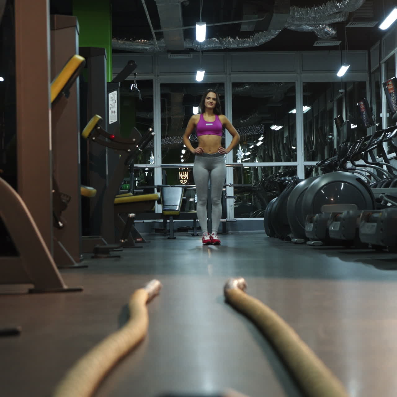 Hermosa mujer en leggings grises se acerca a los cables para hacer ejercicio en el centro deportivo. Una joven delgada y sonriente toma las cuerdas en sus manos. Primer plano.