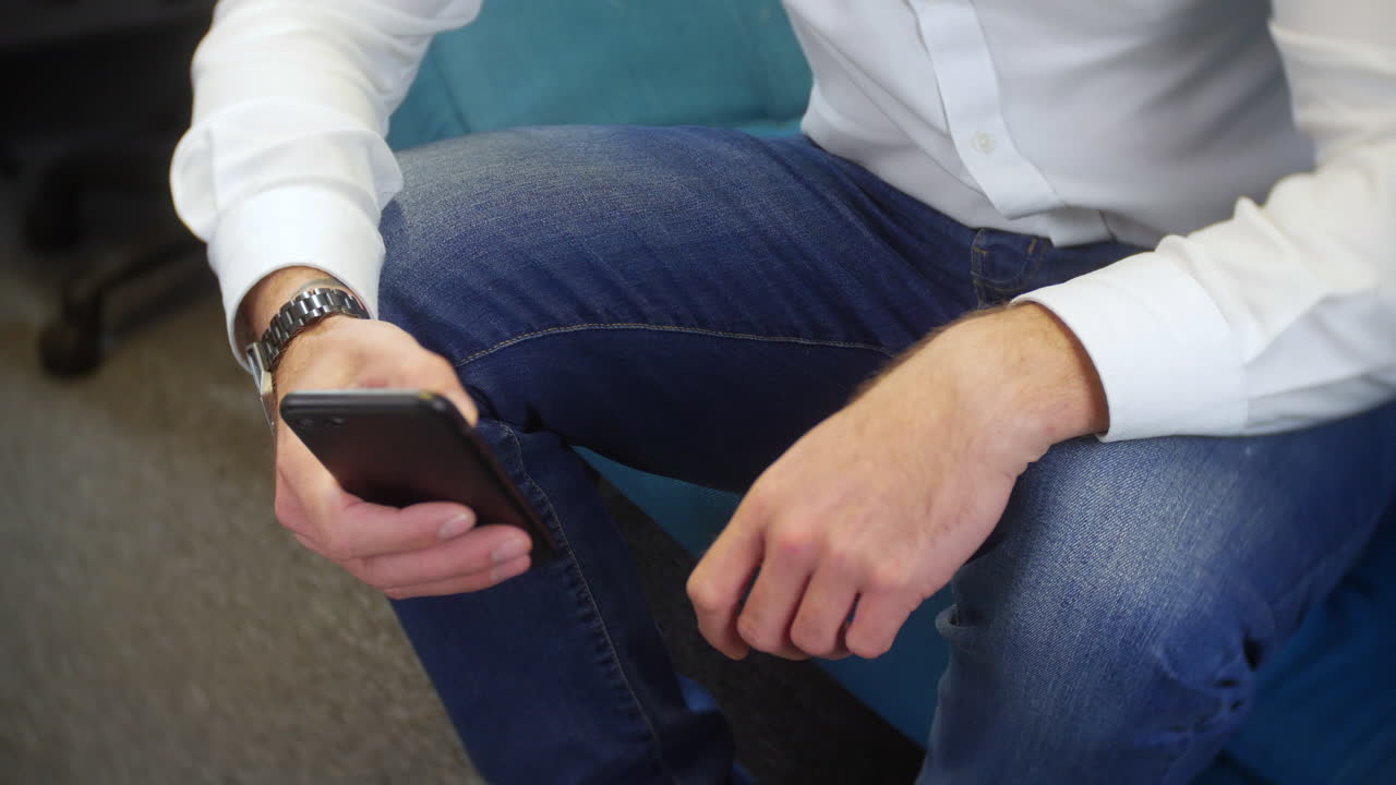 A young man using an app on his mobile cell phone while sitting on a sofa in a home office. The unrecognizable male is wearing jeans and a white shirt