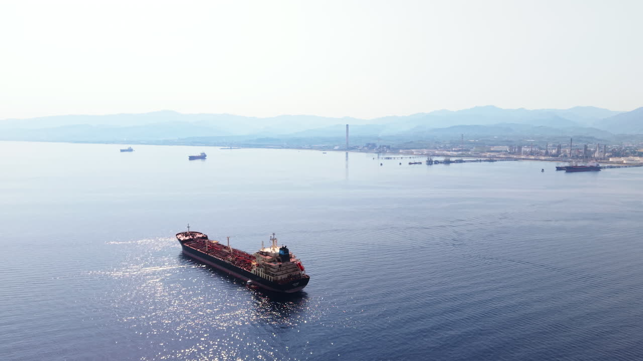 Drone orbits a red tanker ship near an oil refinery in Sicily, with reflections on the calm sea and distant mountains