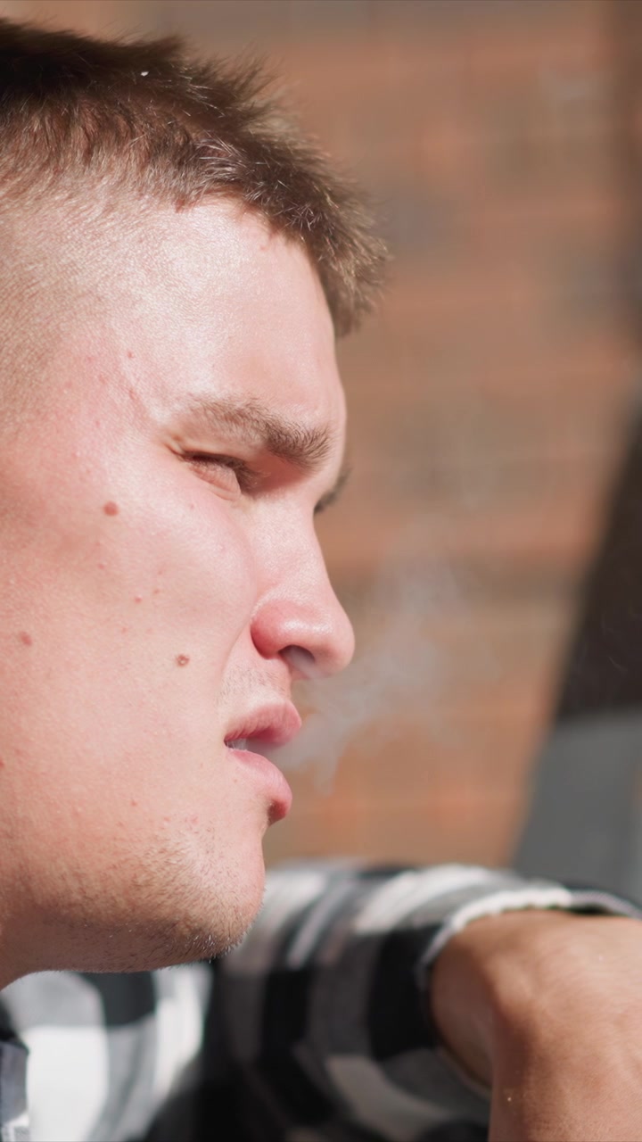 Primo piano di un uomo che fuma, tiene e porta una sigaretta alla bocca. Il soggetto indossa una camicia a quadri con dettagli di texture visibili come i pori della pelle sul viso o le vene della mano. Lo sfondo è sfocato.