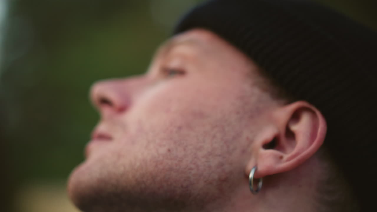 Uomo caucasico che espira il fumo di una sigaretta nella natura, con berretto e orecchino, occhi chiusi e rilassati, calda luce dorata dopo un trekking, atmosfera serena e riflessiva con una delicata texture cinematografica e respiro pacifico