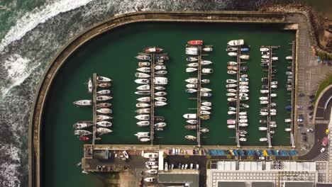 Barco-De-Vela-De-Arriba-Hacia-Abajo-Amarrado-En-El-Puerto,-Vista-De-Pájaro-Del-Ojo-De-Drones-Del-Puerto-De-Hormigón-Con-Olas-Rompiendo