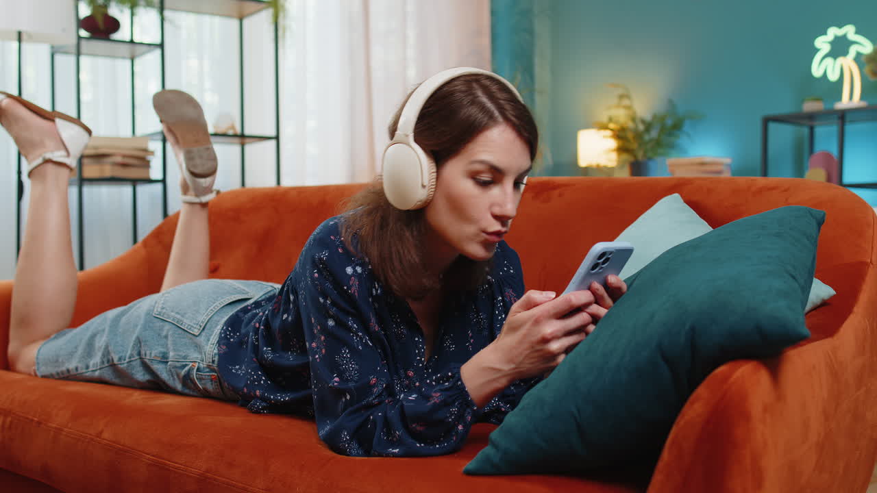 Giovane donna con le cuffie wireless che si rilassa sdraiata sul divano a casa usando lo smartphone ascoltando musica
