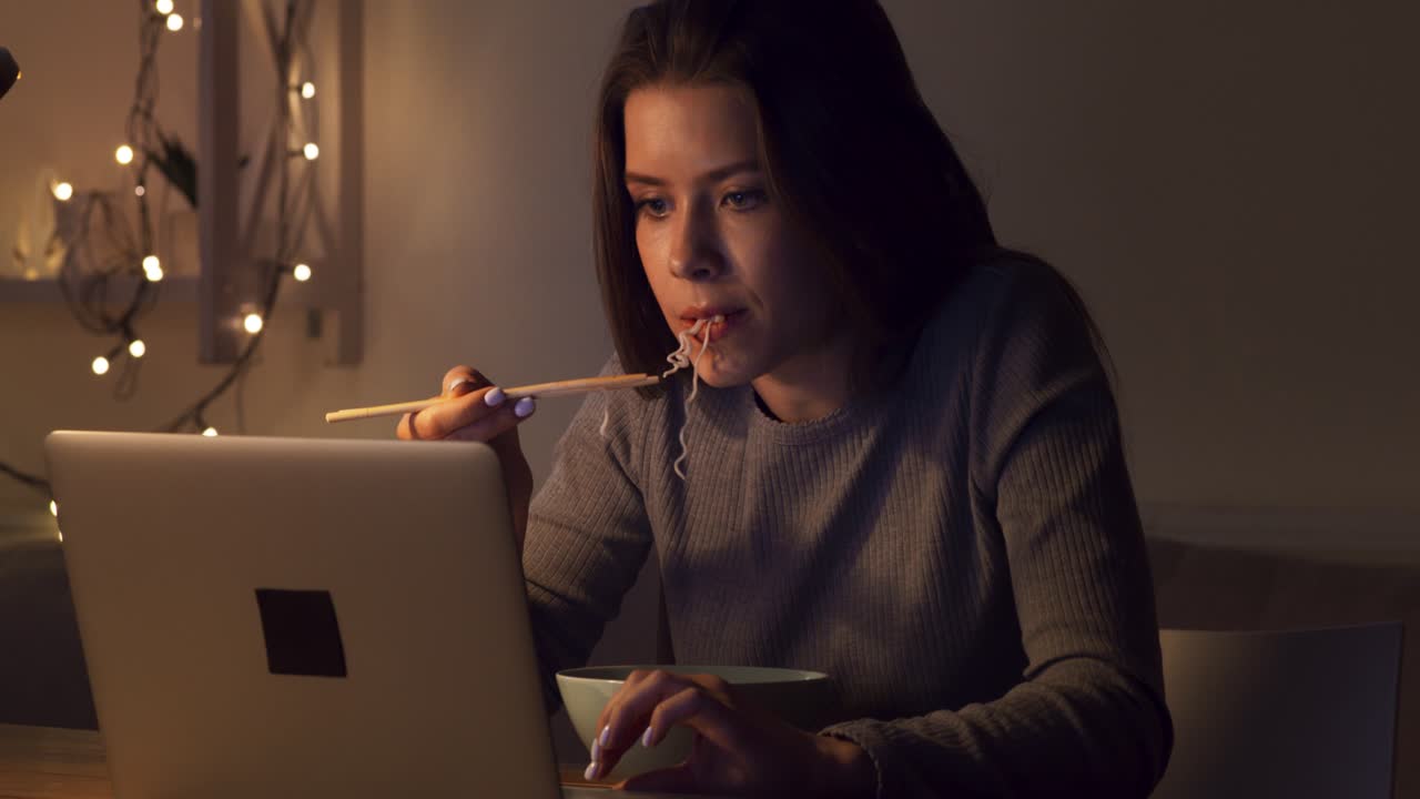 Woman Eating Noodles While Working on Laptop at Night