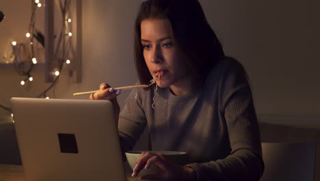woman eating noodles while working on laptop at night