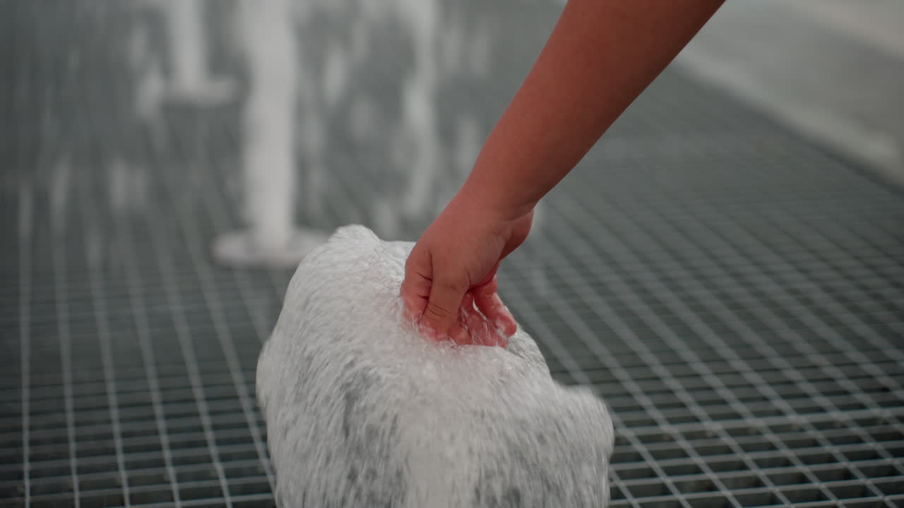 Close view of little girl hand touching bubbling fountain water jet on metal grate, water splashing upward, summer curiosity driving gentle interaction, blurred park background