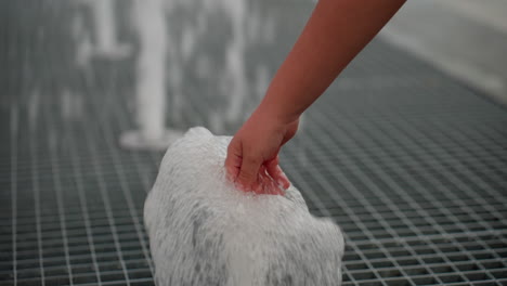 close view of little girl hand touching bubbling fountain water jet on metal grate, water splashing upward, summer curiosity driving gentle interaction, blurred park background
