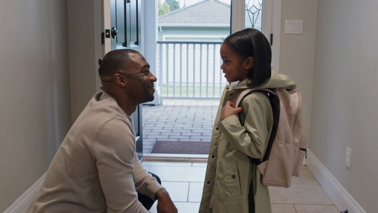 Smoothing father kneeling checking girl's hair, adjusting green coat and pink backpack for school