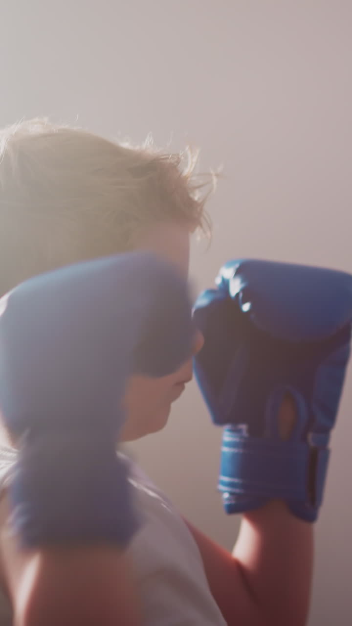 Concentrated little boy in gloves punches bag hanging in gym by rope ladder. Cute child develops professional boxing skills at class at bright light slow motion