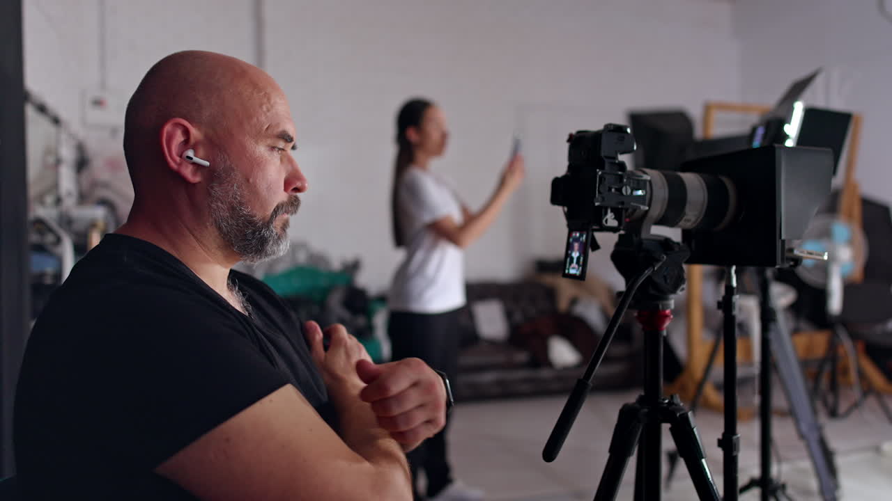 Thoughtful bearded bald man looks at the display of his camera set on tripod. Woman at blurred backdrop uses a smartphone recording a video.