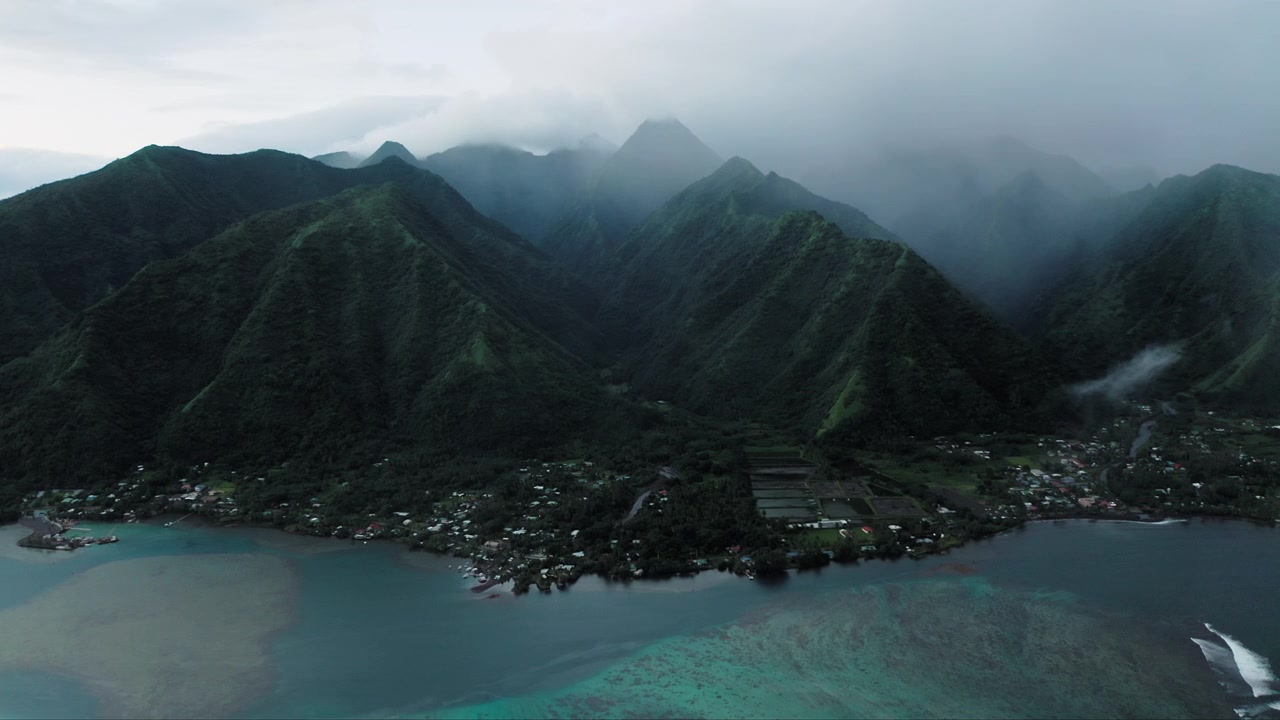 Drone, sisli Teahupoo vadisi, Tahiti üzerinde istikrarlı bir şekilde yükseliyor; dağlar bulutlara karışırken çekim ötesindeki turkuaz kıyı şeridine açılıyor.