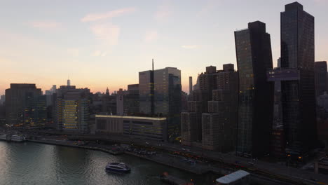 Aufsteigende-Aufnahmen-Von-Modernen-Hochhäusern-Und-Belebten-Mehrspurigen-Straßen-Am-Wasser.-Blick-Gegen-Den-Farbenprächtigen-Sonnenunterganghimmel.-Manhattan,-New-York-City,-Vereinigte-Staaten