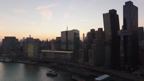 Ascending-footage-of-modern-high-rise-buildings-and-busy-multilane-road-at-waterfront.-View-against-colourful-sunset-sky.-Manhattan,-New-York-City,-USA