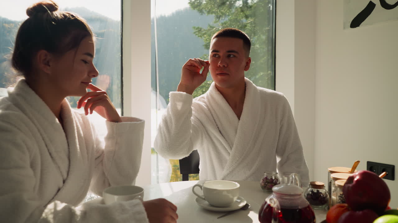 남자가 테이블에 앉아 있는 여자 에 있는 컵에 차 꽃잎을 떨어뜨리고, 남자는 아침에 함께 아침식사하는 뜨거운 차를 즐기며 재미있는 순간을 만니다.
