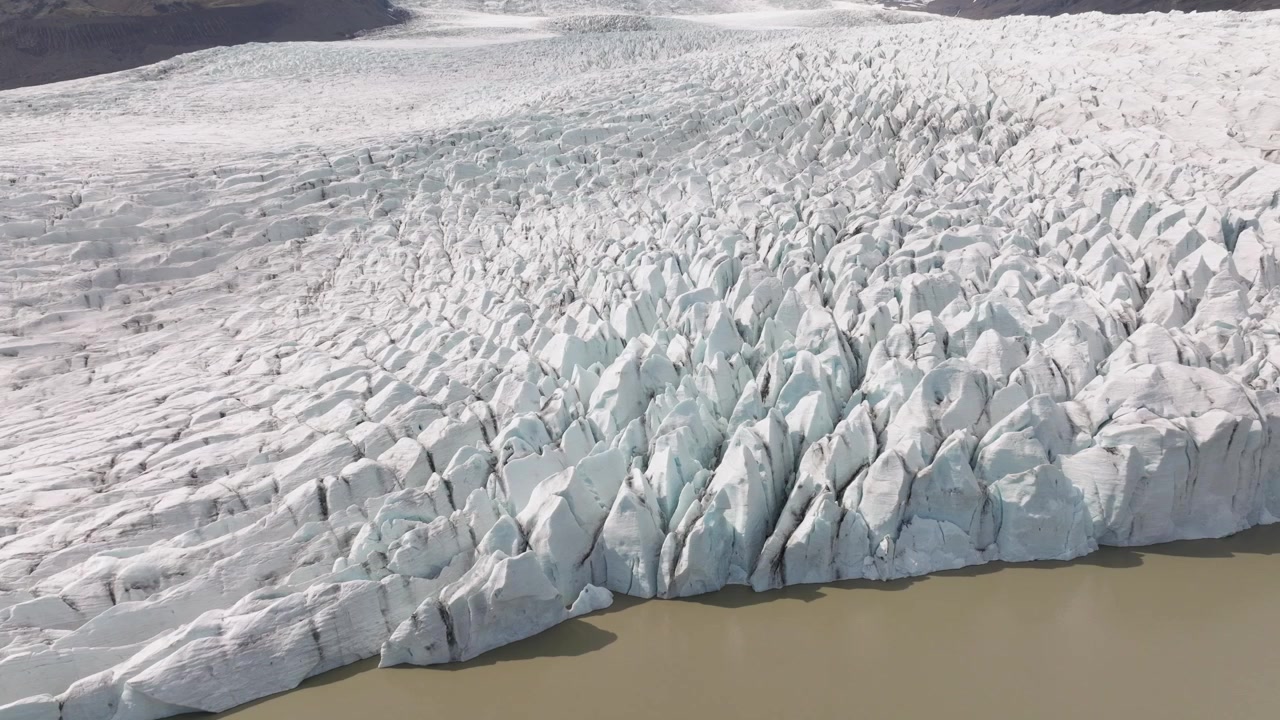 Vista aerea del vasto ghiacciaio Fjallsjökull che scende verso la laguna di Fjallsarlon, Islanda, con profonde fenditure e le aspre vette circostanti sotto un cielo sereno.