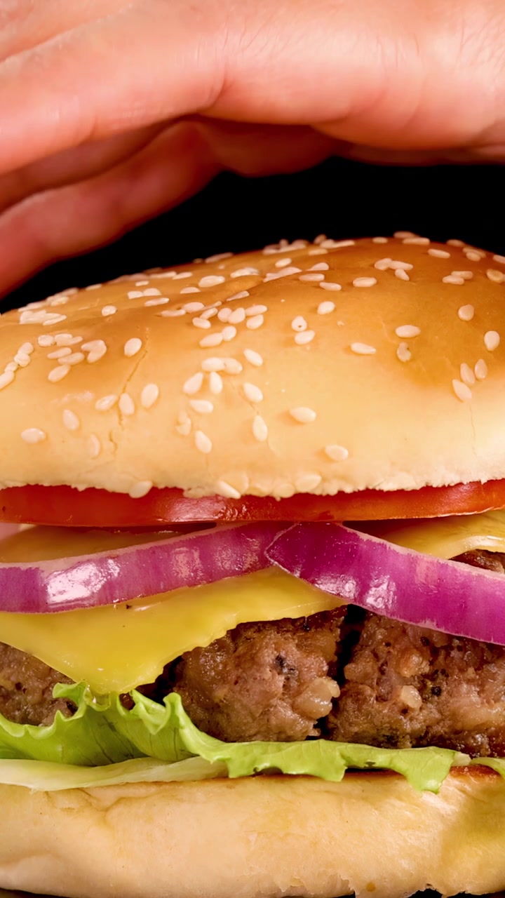 Una mano coloca suavemente un pan de sésamo sobre una hamburguesa de carne con lechuga, queso, tomate y cebolla, bajo una brillante iluminación de estudio, sobre un fondo negro