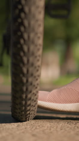 close-up of individual legs in pink sneakers kicking a bicycle tire to check air pressure, lush greenery and trees fill the background, with an indistinct figure moving
