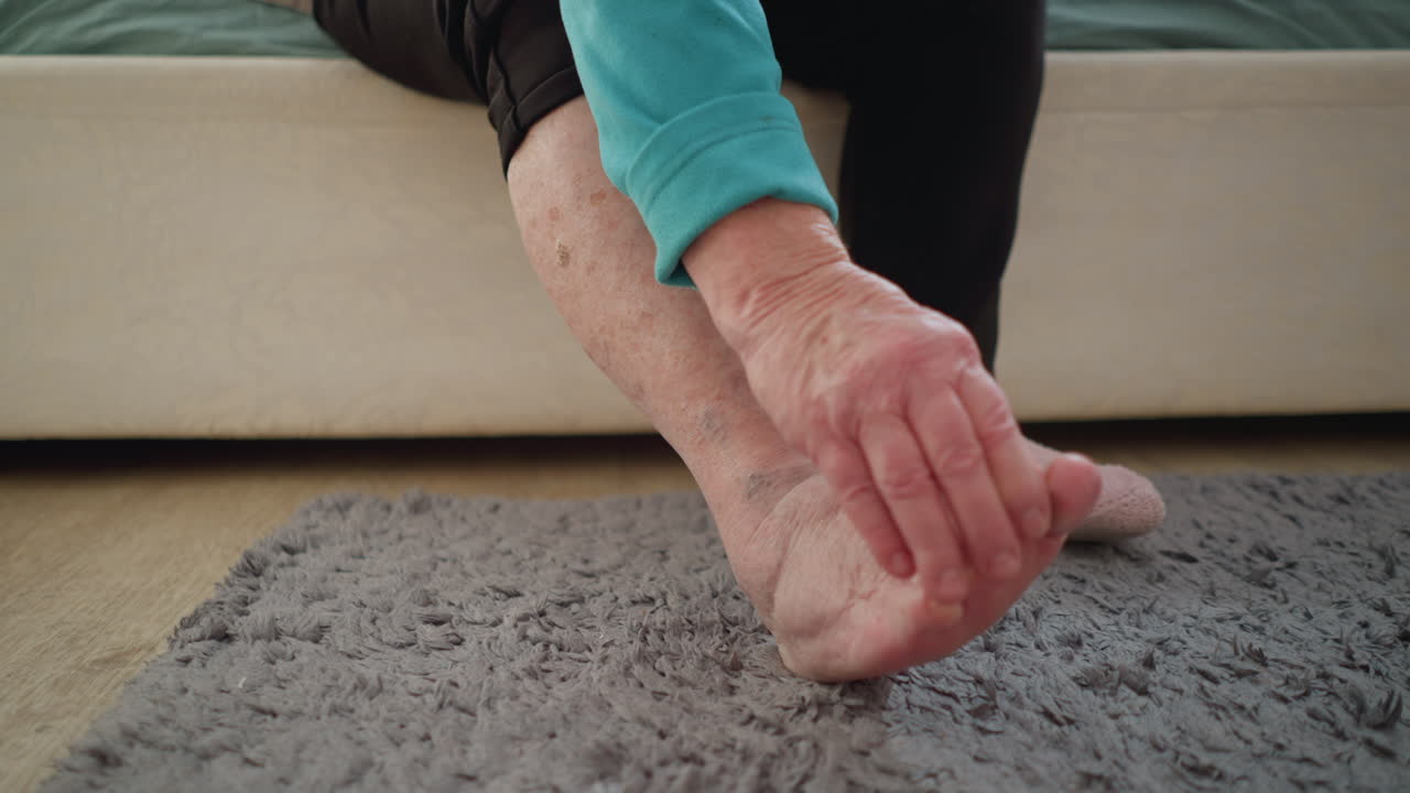 Old woman in green sitting on bed, reaching down to touch her foot. One sock off, the other on. A moment of self-care and comfort in a cozy bedroom, with soft light filling the room