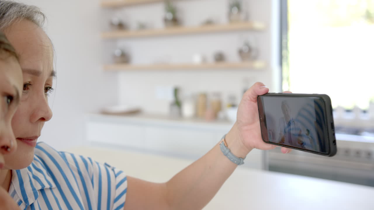 Taking selfie, asian grandmother and child bonding at home in kitchen