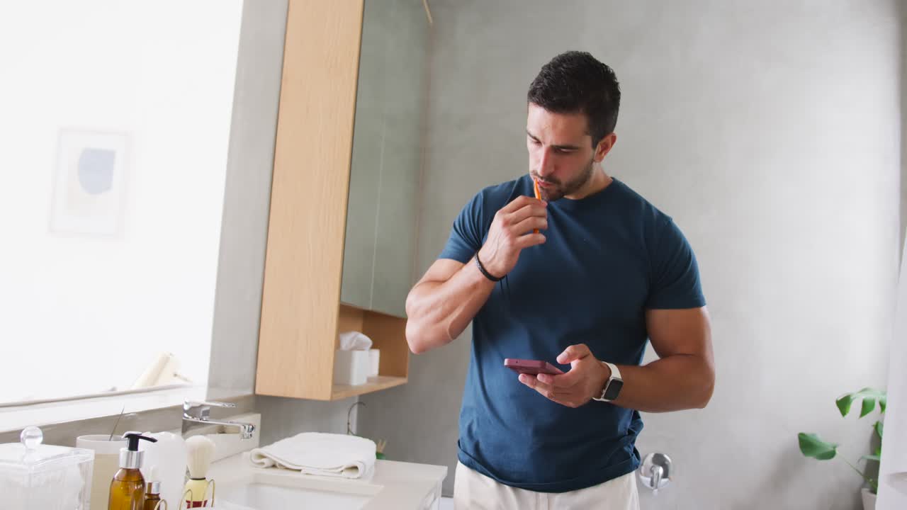 Uomo adulto in piedi al lavandino vicino alla finestra, controlla lo smartphone e poi si lava i denti con uno spazzolino arancione.