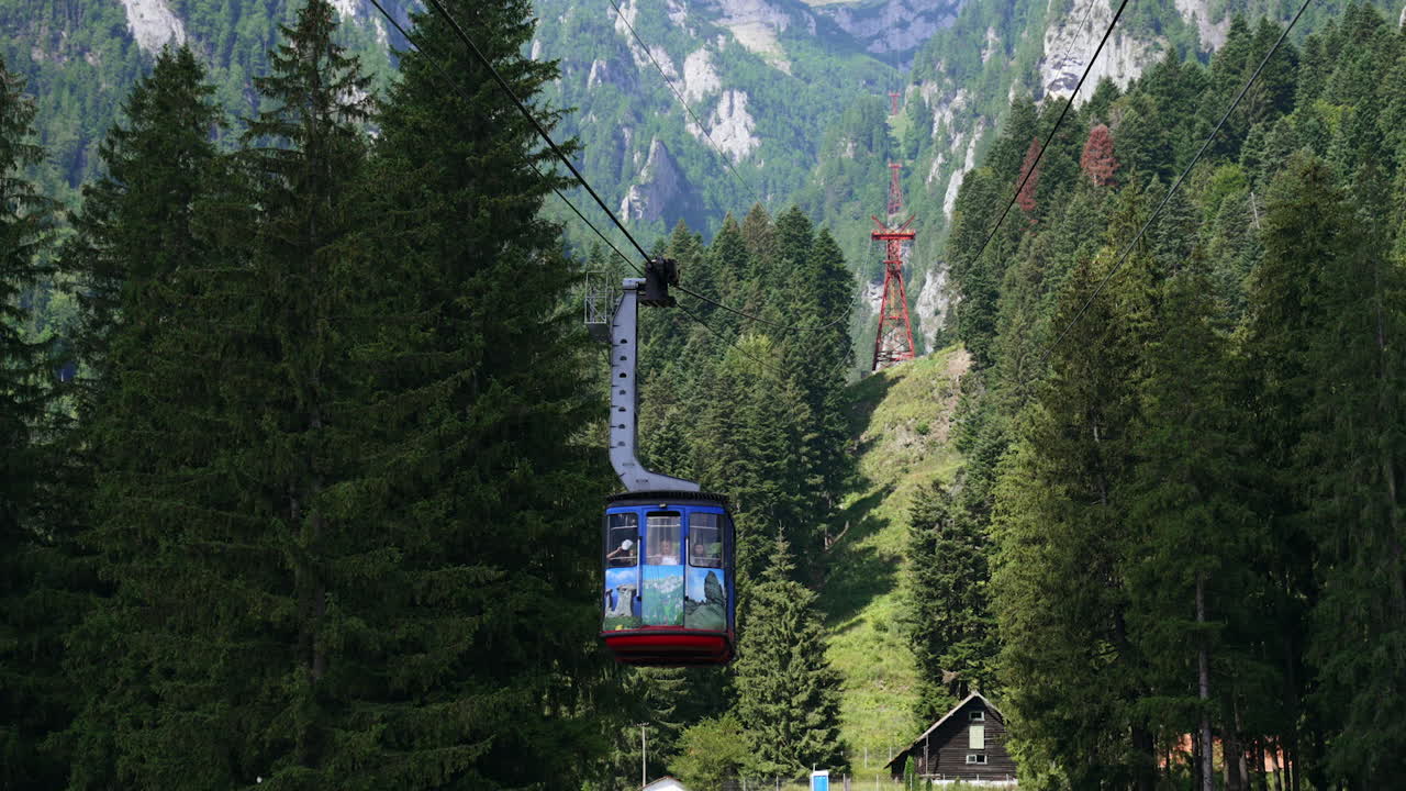 Mountain gondola ride over lush forest. A gondola travels above a dense green forest, leading to majestic mountain peaks in the background on a sunny day