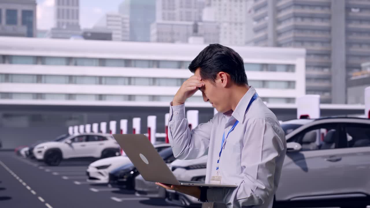 ノートパソコンを使用しているアジア人男性のプロフェッショナルワーカーの側面図。現代の都市ステーションで電気自動車が充電されており、彼は落胆して頷いている。