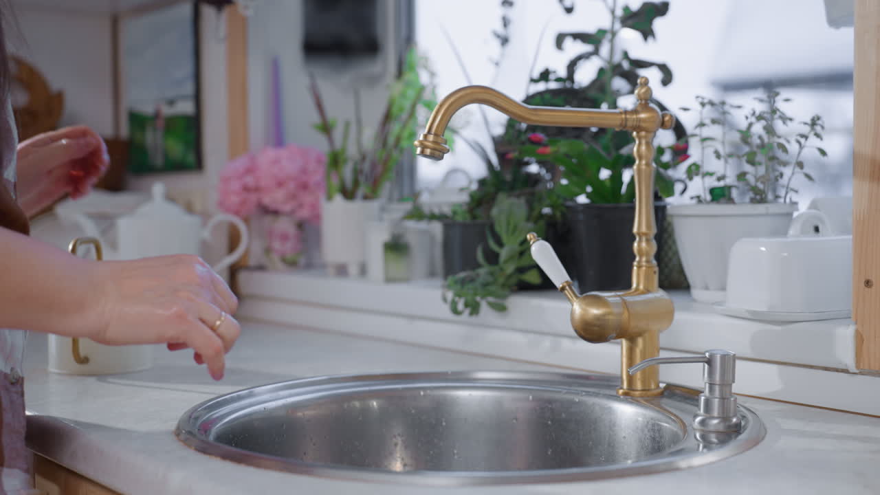 Urban gardener washing hands under rushing tap in modern sink with water flowing over skin, wearing plaid shirt, surrounded by potted plants on ledge, creating serene indoor gardening scene