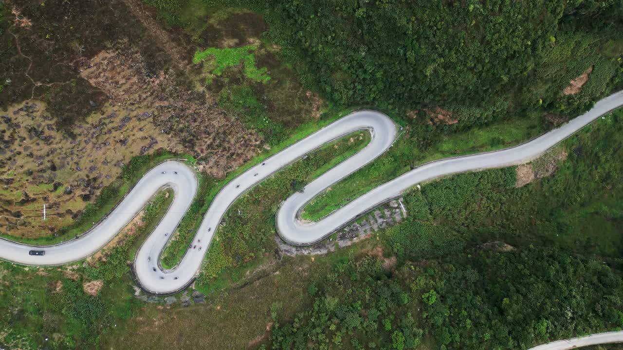 Riprese aeree catturano una stretta strada di montagna con curve strette e ampie che attraversa ripidi pendii verdi, mentre i veicoli si muovono attraverso un paesaggio drammatico negli altipiani del Vietnam settentrionale.