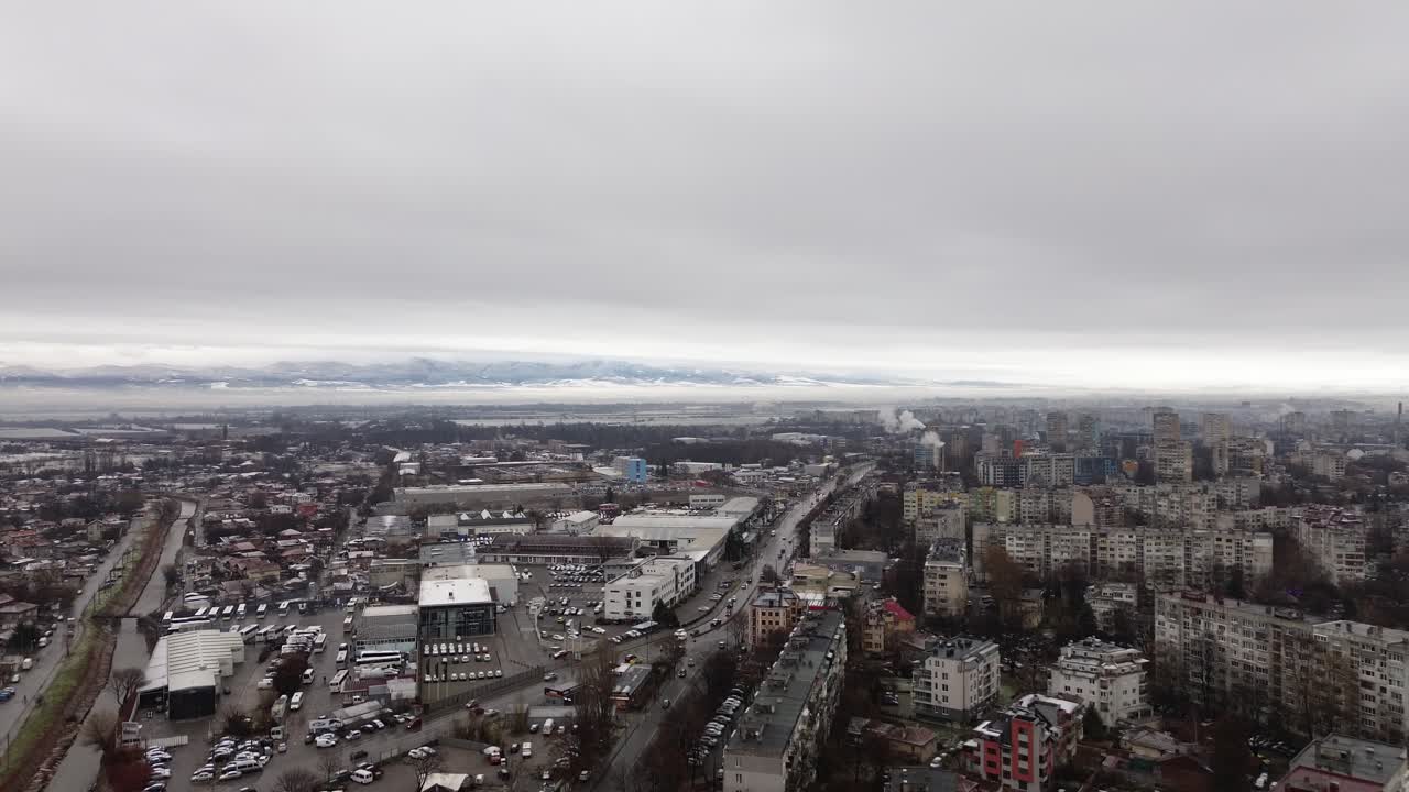 Drone footage over Sofia, Bulgaria, capturing communist-era apartment buildings on the right, a small boulevard with cars, and an industrial district on the left under an overcast winter sky.