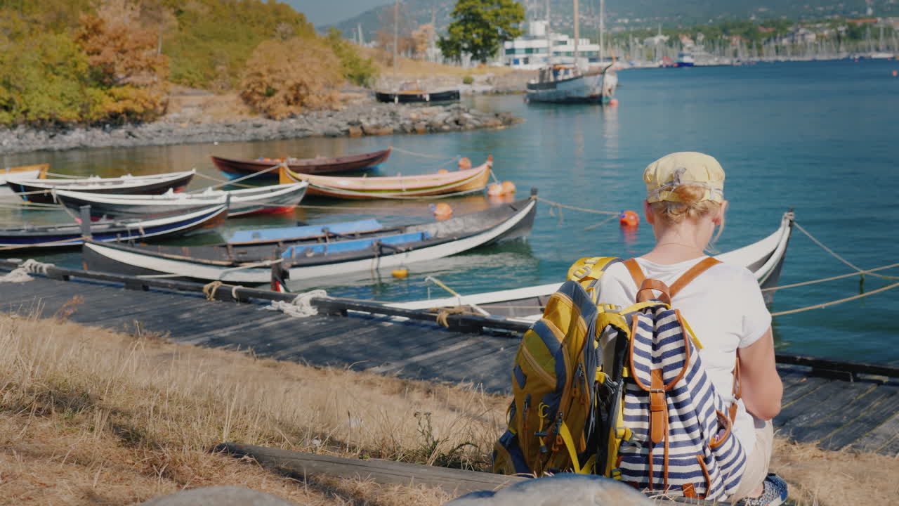 배낭을 메고 있는 한 여성이 어선 근처 노르웨이 오슬로 시 해안가에 앉아 있다