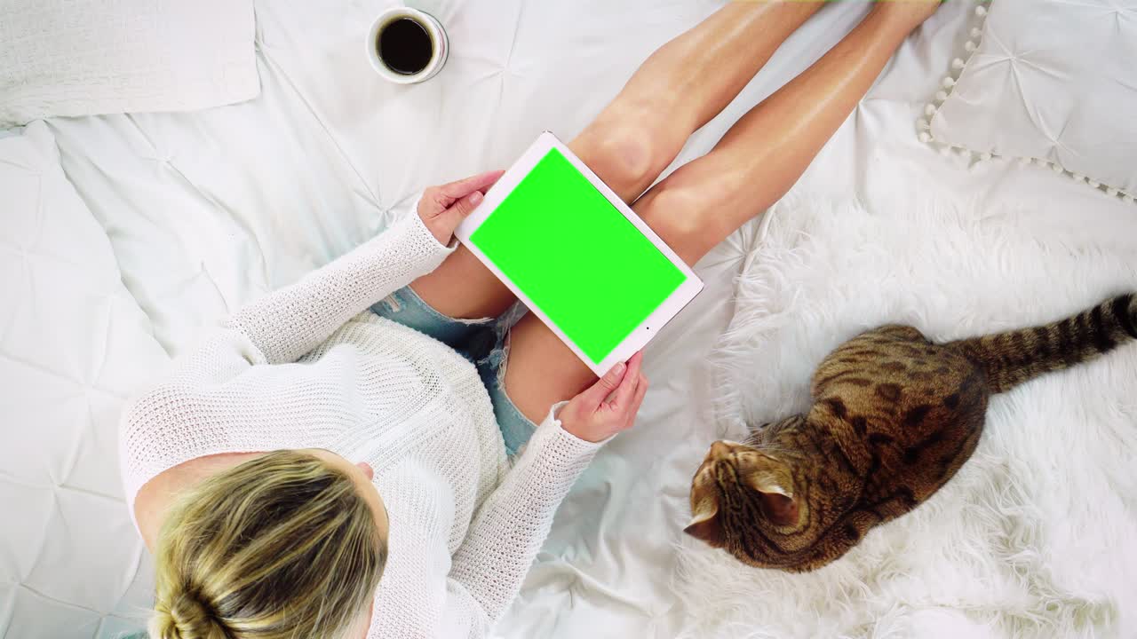 vista de arriba hacia abajo de una mujer sentada en la cama usando un dispositivo de tableta con una pantalla verde en blanco
