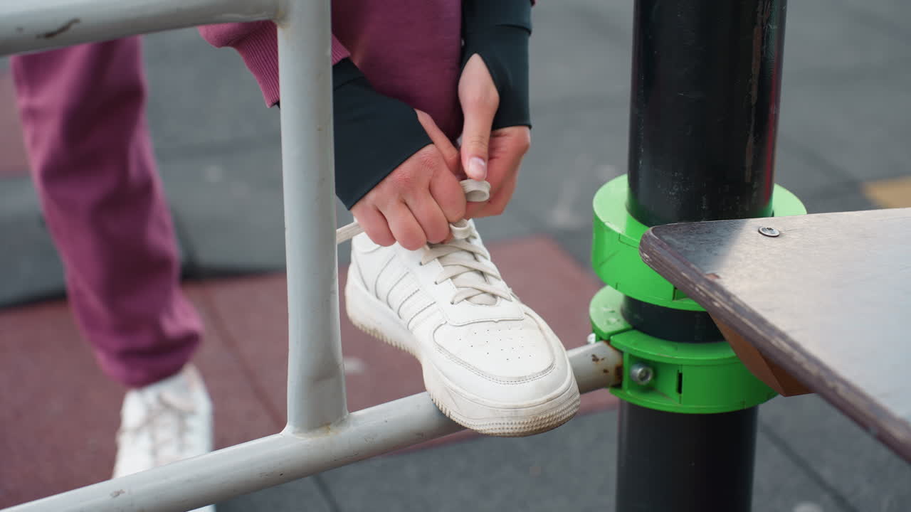 Sporty woman boots perched on iron bar tying canvas sneaker laces with careful focus before outdoor calisthenics session in urban fitness park under open sky near modern apartments