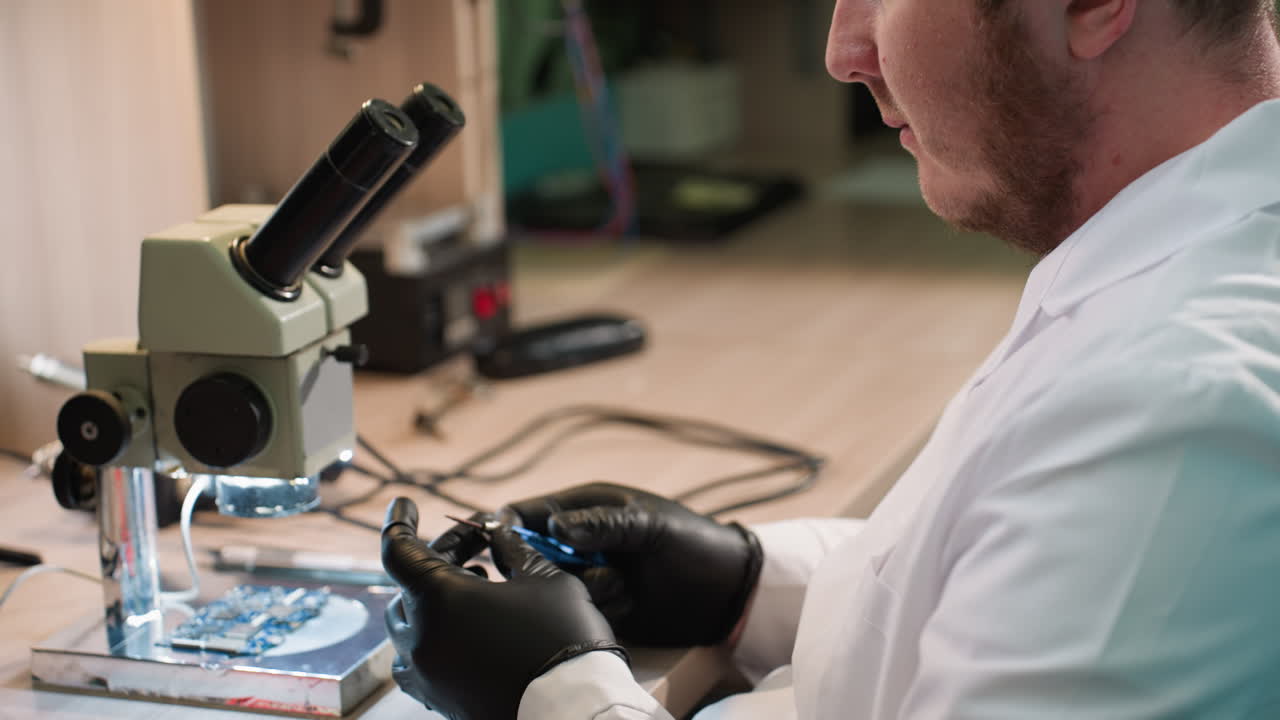 близкий вид техника в лабораторном пальто, сосредоточенного на винте в руке с электронным компонентом под микроскопом и различными инструментами на столе и полке