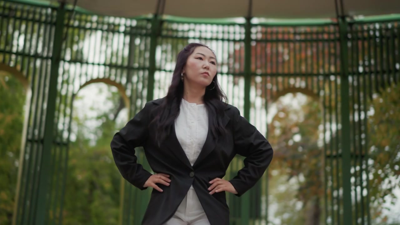 Close-up de uma mulher realizando movimentos de dança em um gazebo ao ar livre com ferragens verdes, vestindo um blazer preto e roupas brancas. Suas mãos movem-se expressivamente enquanto a mulher dança, em um cenário suavemente desfocado.