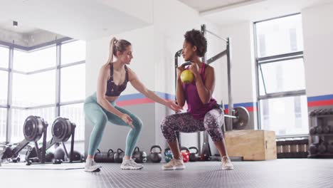 video of diverse female fitness trainer instructing woman doing squats with kettlebell weight at gym