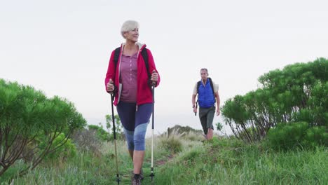 senior hiker couple with backpacks and hiking poles while walking in the grass field.