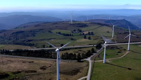 wind-turbines-spinning-in-the-mountains-with-cattle-grazing-nearby,-road-with-a-traveling-car,-sunny-day-and-a-clear-blue-sky