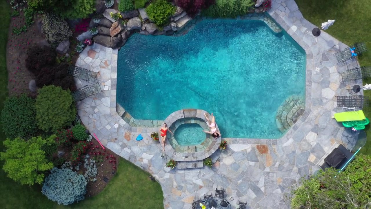two beautiful women of mixed races, lounging by a jacuzzi, within a pool on a beautiful summer day in Connecticut. The camera lifts up slowly, looking straight down on them, with a bird's eye view.
