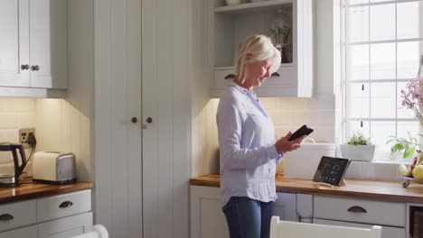 una mujer caucásica senior feliz de pie en la cocina, usando un teléfono inteligente y una tableta.