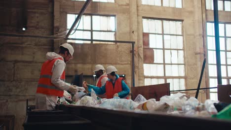 Ein-Team-Aus-Drei-Recyclinganlagenarbeitern-In-Weißen-Uniformen-Und-Orangefarbenen-Westen-Ordnet-Flaschen-In-Einer-Großen-Alten-Recyclinganlage-Nach-Der-Farbe-Des-Kunststoffs