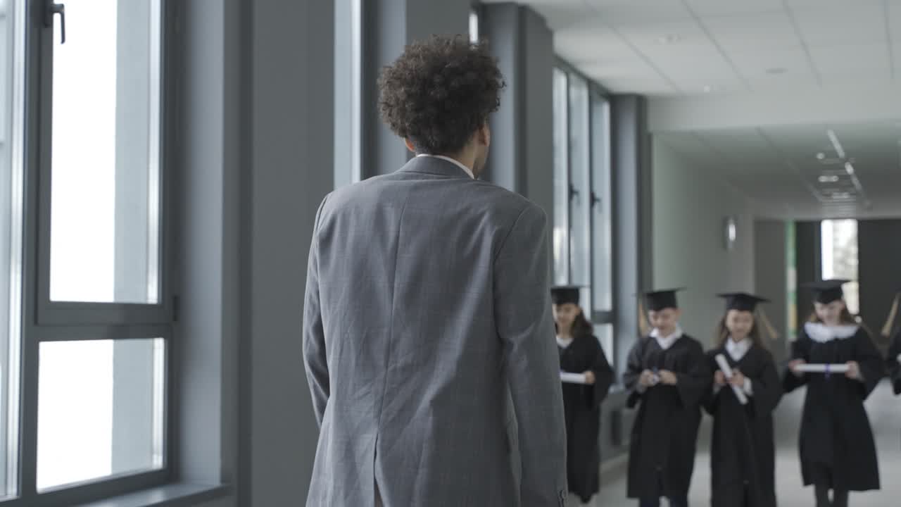 grupo de felices estudiantes de preescolar multirraciales con birrete y bata saludando a su maestra.