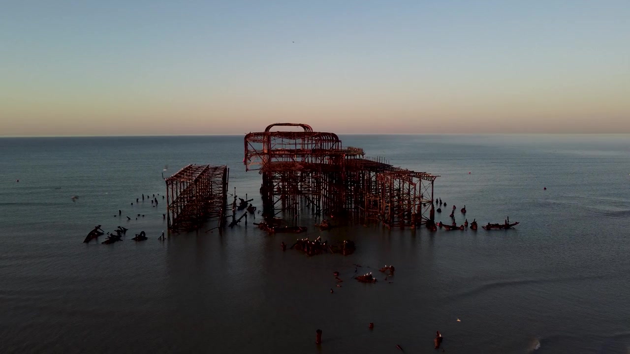 Rotational drone shot of the ruins of the West Pier in Brighton as birds fly past close to the shot and sun rises over the ocean