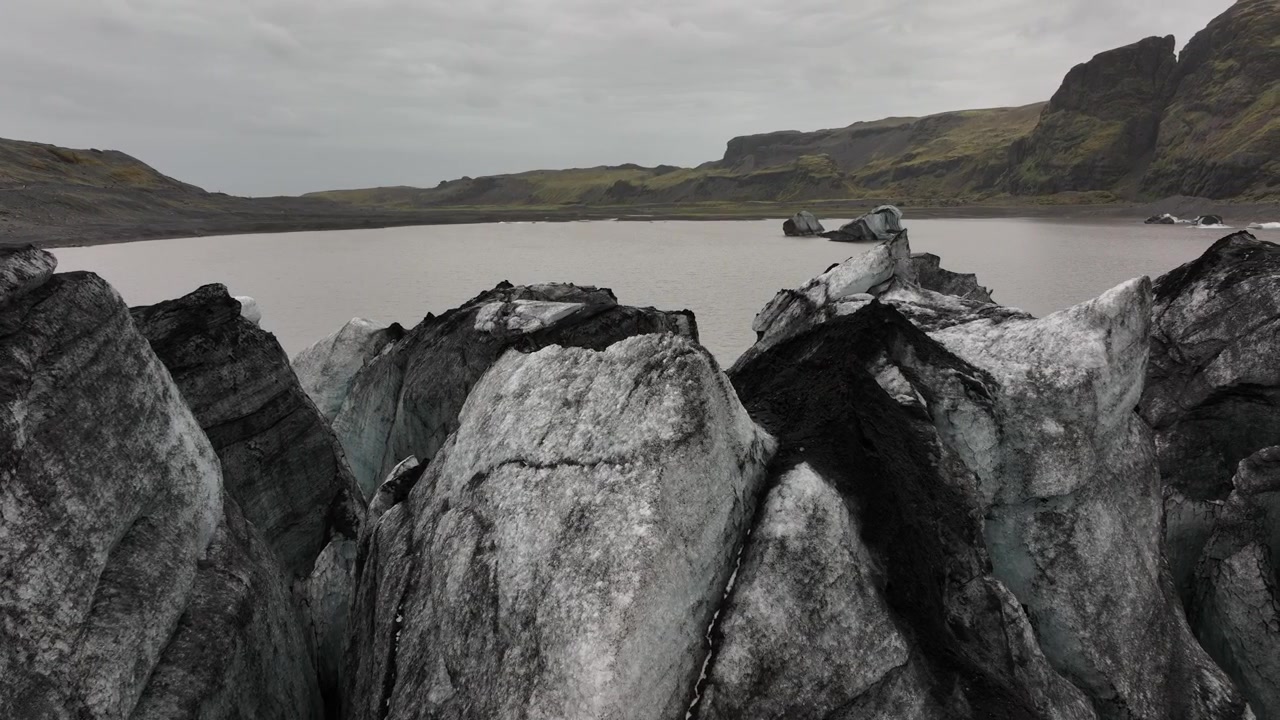 Yksityiskohtainen näkymä terävistä, rosoisista jäämuodostelmista Sólheimajökull-jäätikön reunalla, joka kohtaa jäätikkölaguunin Islannissa, taustalla karua maastoa.