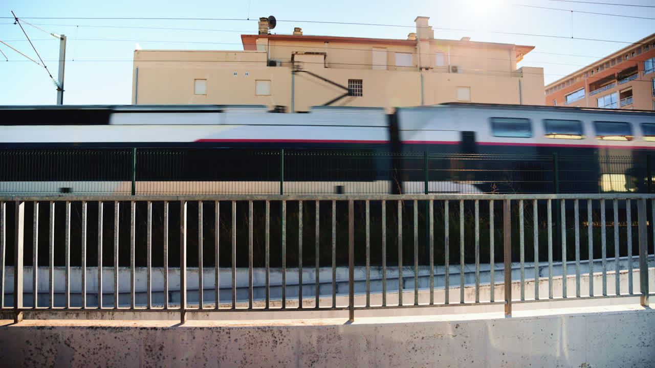 A fast moving passenger train travels past a residential building on a sunny day, captured from behind a metal railing
