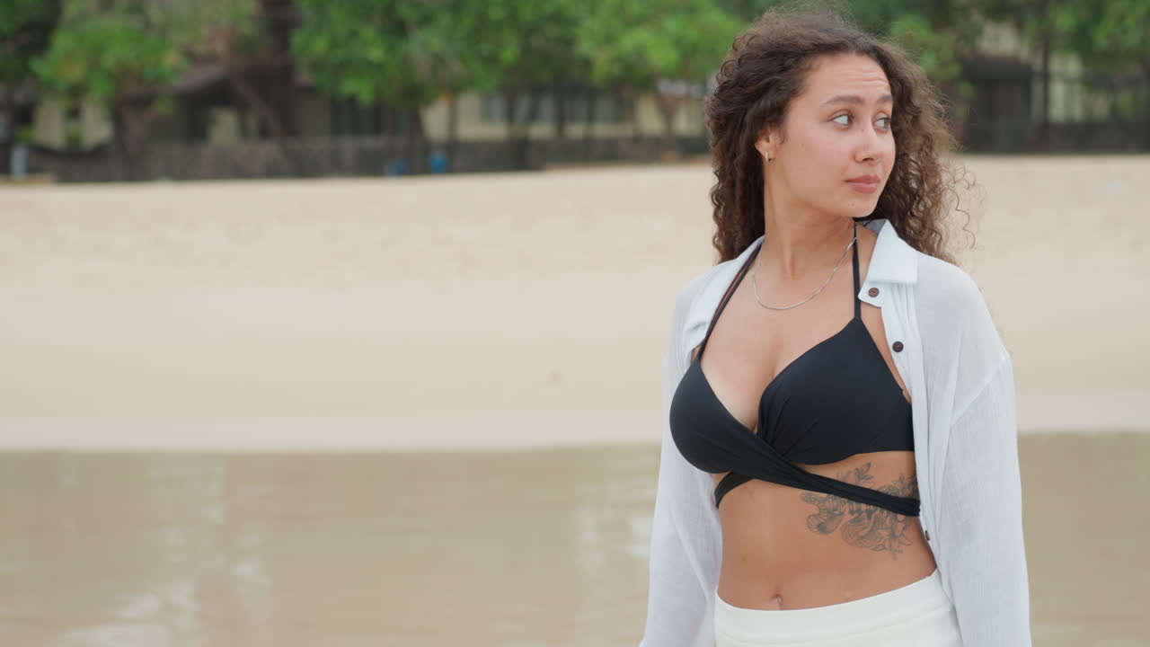 Donna birazziale che passeggia sulla sabbia dell'isola, litorale affollato con bandiere, tatuaggio visibile sulle costole, copricostume bianco sopra un bikini nero, sguardi curiosi, attività in riva al mare, energia positiva e sicura di sé, spontaneo.