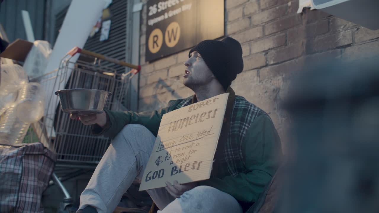 Persona sin hogar sosteniendo cuenco para donaciones y cartel en calle urbana.