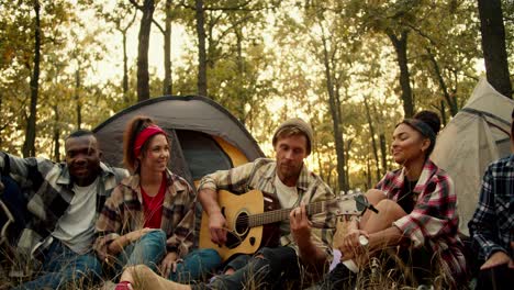 Un-Hombre-Rubio-Toca-La-Guitarra-Mientras-El-Resto-De-Excursionistas,-Durante-Una-Parada,-Cantan-Con-él-Y-Escuchan-Su-Agradable-Música-En-La-Guitarra-Con-El-Telón-De-Fondo-De-Tiendas-De-Campaña-En-Un-Soleado-Bosque-Verde-De-Verano.